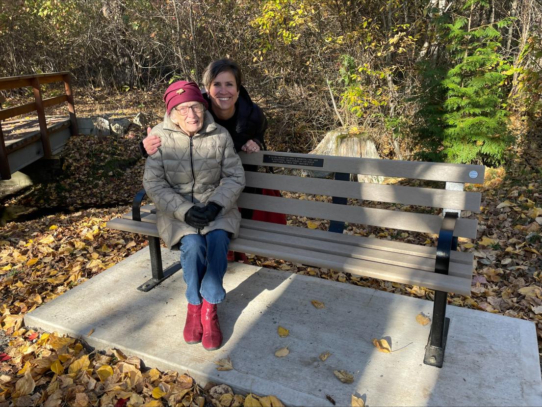 A photo of Evelyn Pousette seated on a memorial bench for her husband, with daughter Ruth behind her
